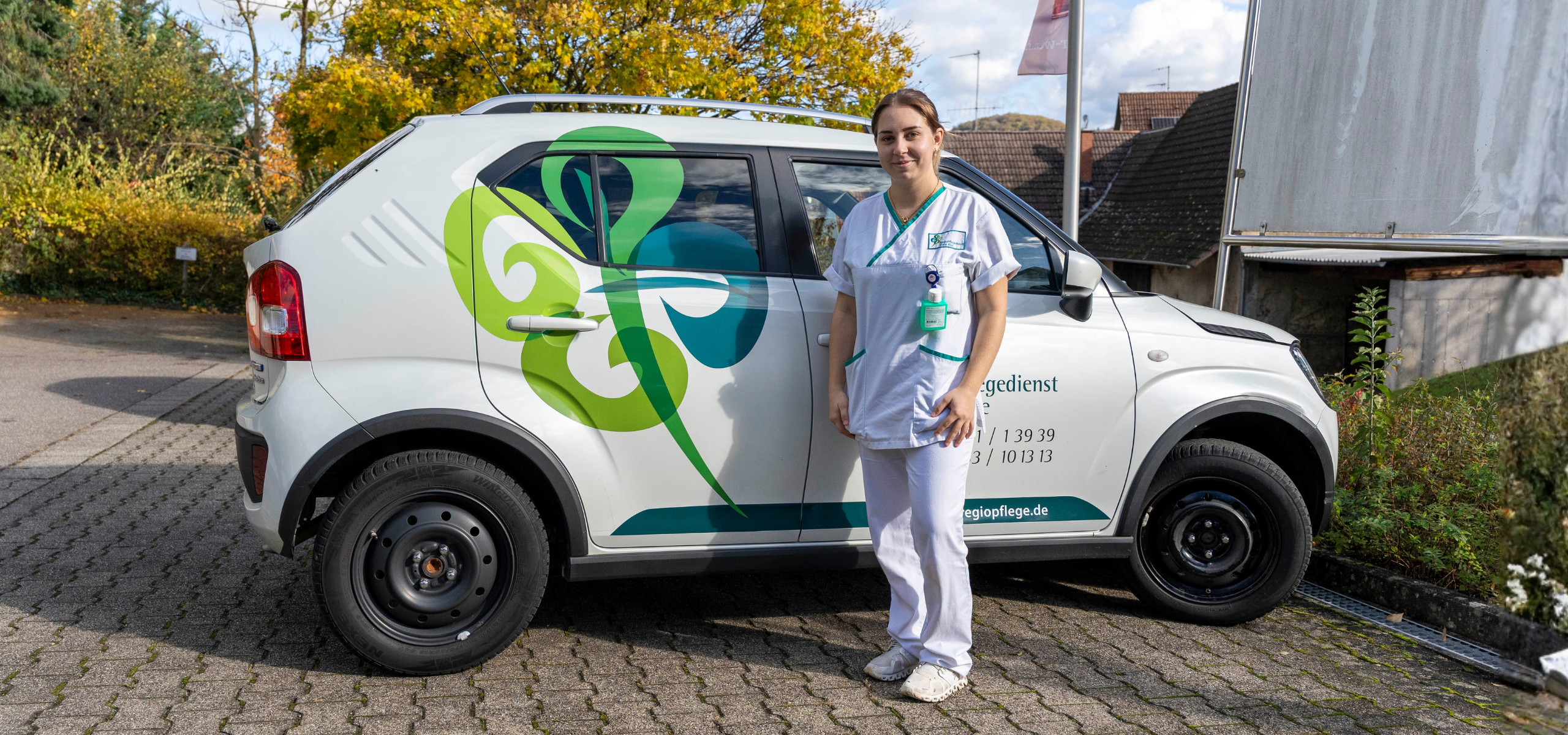 Pflegekraft in Dienstkleidung steht neben einem Dienstfahrzeug des RegioPflegedienst Klaus Klee.